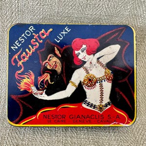 May include: A vintage tin box with a colorful illustration of a woman in a red dress and a devil holding a match. The text on the box reads "Nestor Fausta Luxe" and "Nestor Gianaclis S-A Le Caire - Geneve - Cavalla".