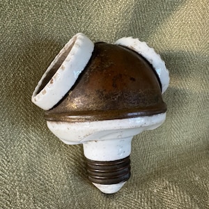 May include: An antique light fixture with a bulb base. The fixture has a copper-colored dome with white ceramic rings and base. The bulb base is a dark brown color. The fixture is on a textured green surface.