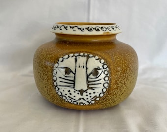 May include: A small, brown ceramic vase with a white stylized cat face design. The vase has a textured surface and a white rim.
