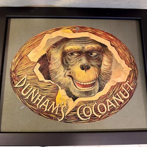 May include: A framed vintage advertisement for Dunham's Cocoanut featuring a smiling monkey's face peeking out of a hole in a coconut shell.
