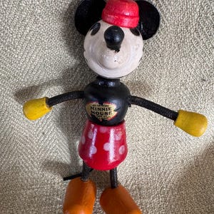 Puede incluir: Mu&ntilde;eca de madera vintage de Minnie Mouse con falda roja de lunares blancos, gorro rojo y guantes amarillos. La mu&ntilde;eca tiene orejas, brazos negros y un torso negro con una etiqueta de Minnie Mouse. Los pies son naranjas.