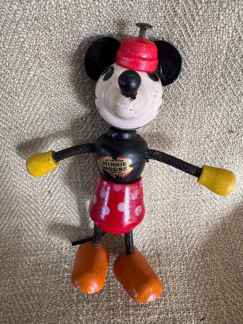 Puede incluir: Mu&ntilde;eca de madera vintage de Minnie Mouse con falda roja de lunares blancos, gorro rojo y guantes amarillos. La mu&ntilde;eca tiene orejas, brazos negros y un torso negro con una etiqueta de Minnie Mouse. Los pies son naranjas.