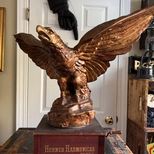 May include: A vintage, gold-painted eagle statue on a wooden music box with the text "Hohner Harmonicas Patent applied for".