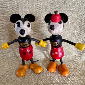 Puede incluir: Dos figuras de madera vintage de Mickey Mouse y Minnie Mouse. Mickey lleva pantalones cortos rojos con botones blancos, guantes amarillos y zapatos naranjas. Minnie lleva una falda roja con lunares blancos y un sombrero rojo. Ambos tienen orejas y brazos negros.