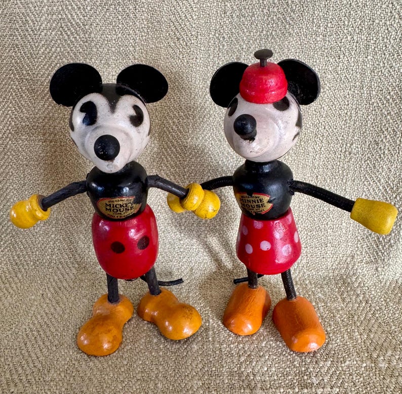 Puede incluir: Dos figuras de madera vintage de Mickey Mouse y Minnie Mouse. Mickey lleva pantalones cortos rojos con botones blancos, guantes amarillos y zapatos naranjas. Minnie lleva una falda roja con lunares blancos y un sombrero rojo. Ambos tienen orejas y brazos negros.