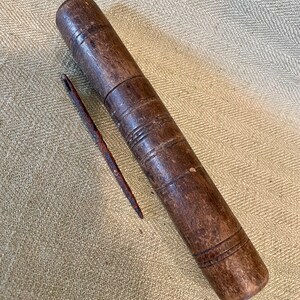 May include: A vintage wooden needle case with a matching needle. The cylindrical case is dark brown with carved horizontal lines. The needle is long and rusty. The items are on a textured, light brown surface.