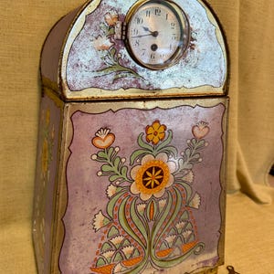 May include: Antique metal clock with a floral design in shades of purple, orange, and green. The clock face is round with a glass cover. A small key rests beside the clock. The clock is approximately 8 inches tall.
