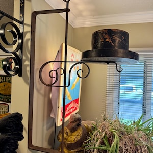 May include: An antique, wrought iron sign holder with a heart-shaped finial. A black top hat rests on a shelf attached to the sign holder. The sign holder is mounted on a beige wall, with a vintage Cosmopolitan sign visible.