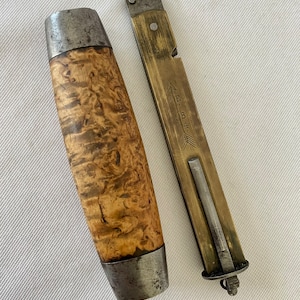 May include: An antique knife with a wooden handle and a metal blade. The handle is light brown with a unique wood grain pattern. The metal blade is housed in a brass sheath. The sheath has the text "JE 1884" engraved on it.