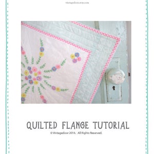 May include: A close-up of a white quilt with a pink and white gingham border. The quilt has a floral design in the center with pink, yellow, and blue flowers. The quilt is lying on a light blue door with a white doorknob. The text "QUILTED FLANGE TUTORIAL" is at the bottom of the image. The text "Vintagedoor 2016. All Rights Reserved." is below the tutorial title.