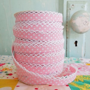 May include: A stack of pink ribbon rolls with white polka dots and a white scalloped lace trim. The ribbon is neatly coiled, showcasing the delicate design. The background includes a vintage door and a colorful patchwork quilt.