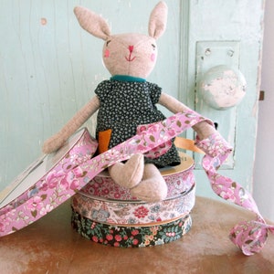 May include: A stack of pink and floral patterned ribbon spools with a plush bunny doll sitting on top. The bunny is wearing a dark blue dress with white polka dots.