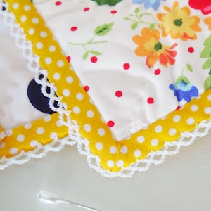 K&ouml;nnte beinhalten: Nahaufnahme eines wei&szlig;en Stoffes mit gelbem Tupfenrand und wei&szlig;er H&auml;kelkante. Der Stoff hat ein Blumenmuster mit roten, gelben und blauen Blumen.