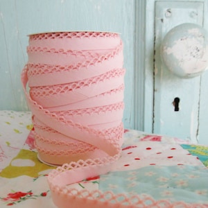 May include: A spool of pink elastic trim with a delicate lace edge. The trim is wound around a white cardboard spool and is sitting on a colorful patchwork fabric.