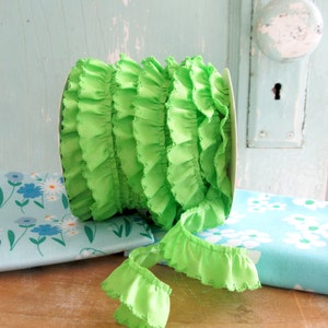 May include: A roll of bright green ruffled trim with a scalloped edge. The trim is on a white spool and is sitting on top of two pieces of blue fabric with white floral patterns.