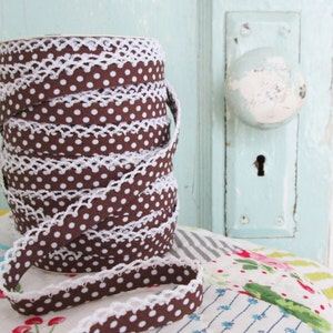May include: Brown and white polka dot ribbon with white lace trim. The ribbon is rolled up and is sitting on a colourful fabric background.
