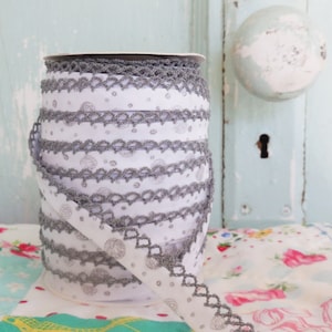 May include: A spool of white and gray trim with a delicate lace pattern. The trim has a repeating pattern of small circles and a scalloped edge.
