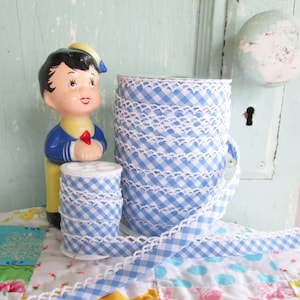 May include: Two spools of blue and white gingham ribbon with white crochet trim. The ribbon is on a colorful patchwork quilt.