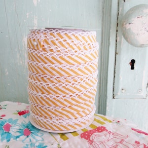 以下が含まれることがあります： 白と黄色のストライプのリボンが巻かれたスプール。白いレースのトリムが付いています。リボンは白いボール紙のスプールに巻かれています。背景は、白いドアノブが付いた水色のドアです。