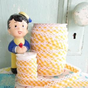 May include: Yellow and white gingham ribbon with white lace trim. The ribbon is wound around a spool and there is a small doll figurine in the background.