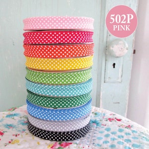 May include: A stack of ten polka dot fabric ribbon rolls in various colors, including pink, red, orange, yellow, green, blue, and black. The ribbon rolls are stacked on a floral patterned surface. The text "502P PINK" is visible in the upper right corner of the image.