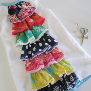 Peut inclure: Une robe blanche sans manches avec une bordure bleue autour de l'encolure et de l'ourlet. La robe pr&eacute;sente un design de volants superpos&eacute;s avec diff&eacute;rents tissus color&eacute;s, notamment des motifs floraux, &agrave; pois et paisley.