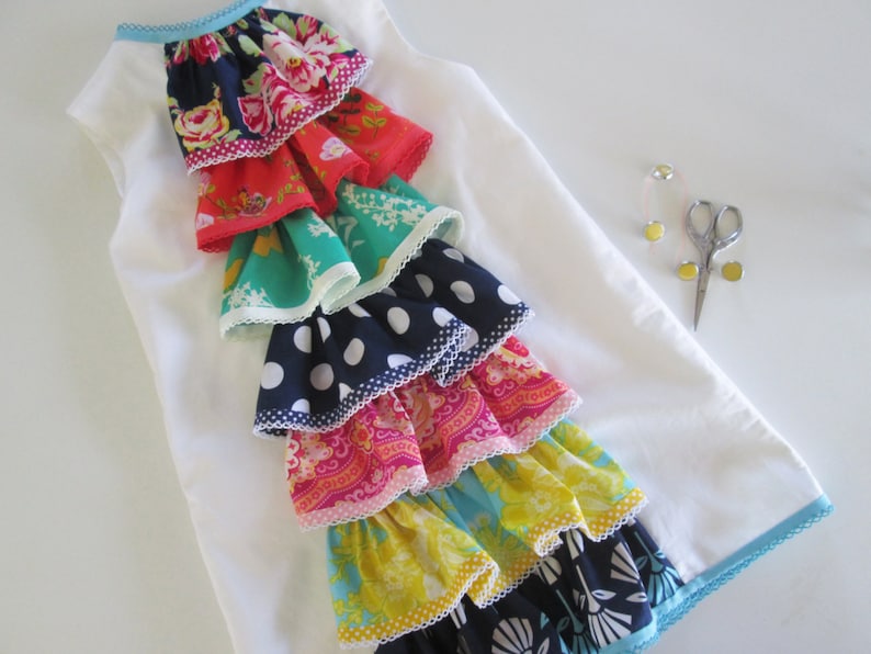 Peut inclure: Une robe blanche sans manches avec une bordure bleue autour de l'encolure et de l'ourlet. La robe pr&eacute;sente un design de volants superpos&eacute;s avec diff&eacute;rents tissus color&eacute;s, notamment des motifs floraux, &agrave; pois et paisley.