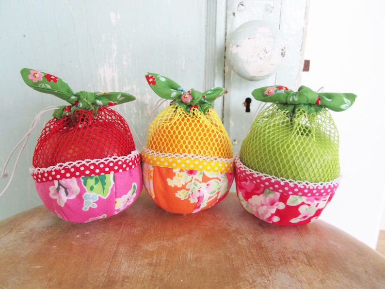 K&ouml;nnte beinhalten: Drei stoffbezogene, fruchtf&ouml;rmige Beutel mit Netzgewebe. Die Beutel sind rot, orange und gr&uuml;n und haben ein Blumenmuster.