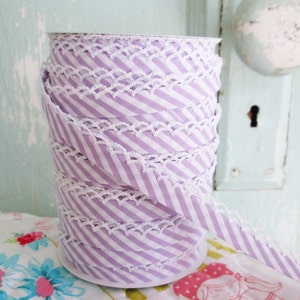 May include: A roll of lavender and white striped ribbon with a white lace trim. The ribbon is on a white spool. The background includes a light blue door and a floral patterned fabric.