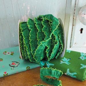 Könnte beinhalten: Eine Spule mit grünem Rüschenbesatz mit einem lindgrünen Rand. Der Besatz befindet sich auf einer weißen Spule und liegt neben zwei Stoffstücken. Ein Stoff ist türkisfarben mit einem Blumenmuster und der andere ist grün mit einem weißen Hirschmuster.