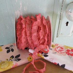May include: A spool of pink ruffled trim with a beaded edge. The trim is on a white background and there is a floral fabric in the background.