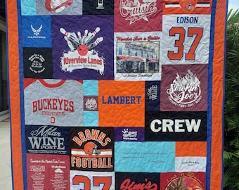 May include: A colorful quilt made from repurposed t-shirts. The quilt features a variety of designs, including sports teams, restaurants, and local businesses. The quilt is a unique and personalized gift for a sports fan or someone who loves their hometown.