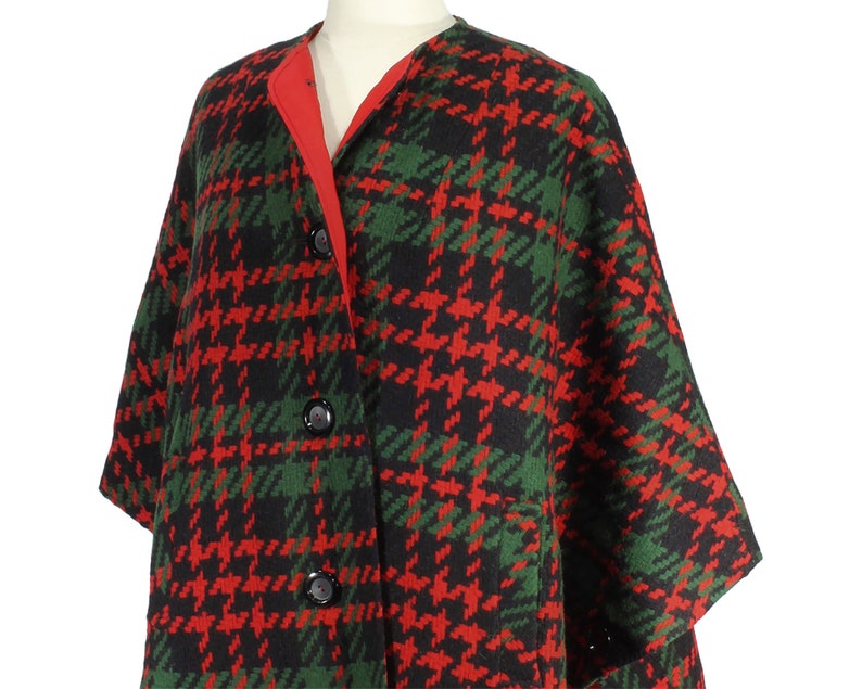 May include: A red and green plaid wool poncho with a black background. The poncho has a red lining and three black buttons down the front.
