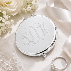 May include: A silver compact mirror with engraved initials, resting on a white satin surface. The mirror is round and has a shiny, reflective surface. A diamond ring and white flowers are also in the image.