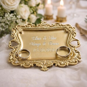 May include: A gold-colored, ornate ring holder with the inscription "Ethan & Chloe, Always & Forever, 05.20.2026." Two wedding rings are placed in the holder. The background includes white flowers and lit candles.