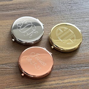 May include: Three round, personalized compact mirrors in silver, gold, and rose gold. The silver mirror has an engraved monogram. The gold mirror features a monogram design. The rose gold mirror is engraved with the name "Jessica" and the date "08-25-25".
