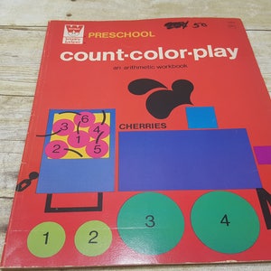 May include: A red preschool workbook titled "count-color-play: an arithmetic workbook" with a colorful illustration of a train carrying cherries. The cherries are numbered 1 through 6. The train cars are numbered 1 through 4.