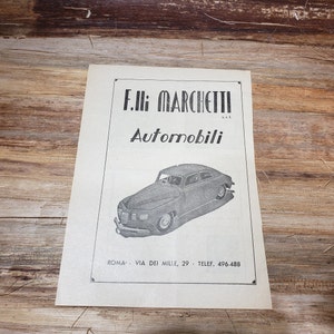 May include: Black and white advertisement for F.lli Marchetti Automobili, a car dealership in Rome, Italy. The ad features a black car and the text "F.lli Marchetti Automobili, s.r.l. Roma: Via dei Mille, 29 - Telef. 496.488"