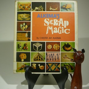 May include: A vintage book titled "Alkema's Scrap Magic" by Chester Jay Alkema, featuring a collage of colorful craft projects. A wooden cat figurine stands in front of the book. The book is displayed on a black stand.