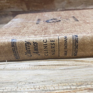 May include: A vintage book titled "Cherry Ames, Clinic Nurse" by Julie Tatham. The book has a textured, tan cover with black lettering and a small illustration. The book is lying on a wooden surface.
