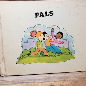 May include: A vintage children's book titled "PALS" with a colorful illustration of three children playing on a green field. The children are wearing different colored clothing and have different skin tones.