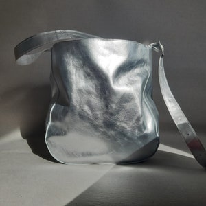 Puede incluir: Un bolso de hombro de cuero plateado con un diseño holgado y sin estructura. El bolso tiene una sola correa y una cremallera.
