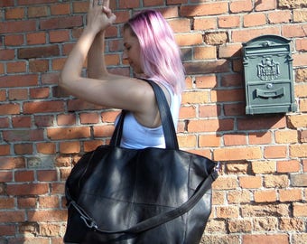 May include: A black leather tote bag with a long strap. The bag has a large compartment and a zippered closure. The bag is being held by a person with pink hair.