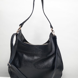 Könnte beinhalten: Schwarze Hobo-Tasche aus Leder mit einem langen Schulterriemen und einem kürzeren Griff. Die Tasche hat einen goldfarbenen Reißverschluss und Beschläge. Das Leder hat ein strukturiertes Aussehen. Die Tasche steht vor einem weißen Hintergrund.