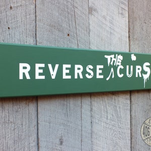 May include: A green wooden sign with white text that reads "The Reverse Curse" and a small horseshoe symbol. The sign is mounted on a wooden wall.