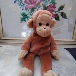 May include: A brown plush monkey toy with a light brown face and black eyes. The monkey is sitting with its legs crossed and its arms hanging down.