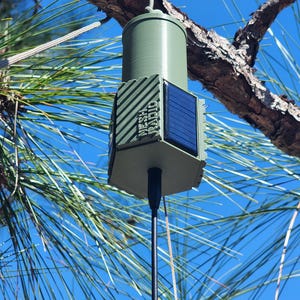 May include: A green cylindrical device with a solar panel and the text "MESH RADIO" is suspended from a tree branch against a blue sky. The device has a black antenna and is likely used for communication.