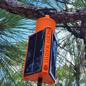 May include: A bright orange triangular mesh radio device featuring black solar panels on its sides and the text MESH RADIO embossed vertically. The device has a textured circular top cap and a black cable connected to its base, hanging from a tree.