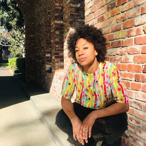 May include: A person with curly dark hair is wearing a colourful short-sleeved shirt with a geometric pattern and black trousers. They are sitting on a concrete step with their legs crossed and looking at the camera.