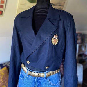 May include: A navy blue cropped blazer with gold buttons and a decorative emblem. The blazer is paired with blue jeans and a gold-toned belt. The jacket is displayed on a black mannequin head.